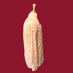 May include: A cream-colored button-down shirt with a geometric pattern of black lines and small red and black squares. The long-sleeved shirt is displayed on a mannequin against a dark red background.
