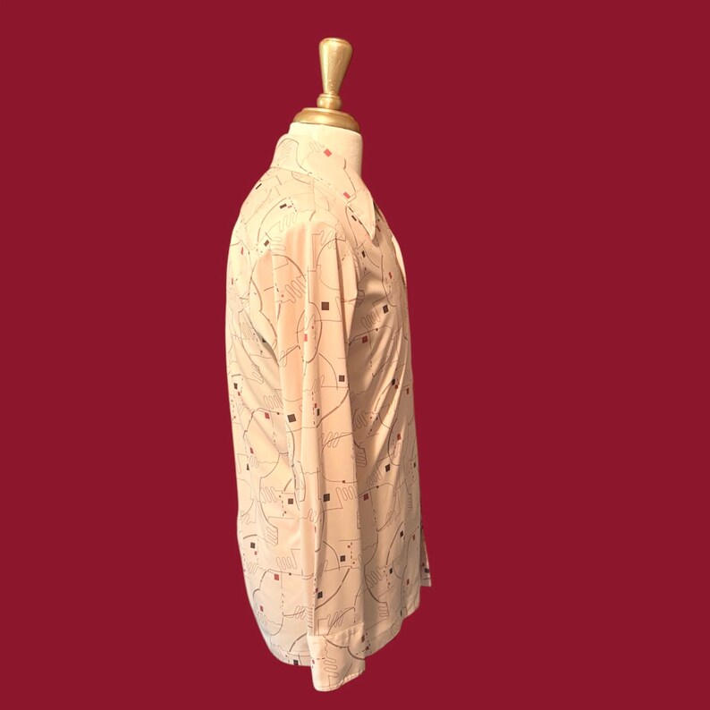 May include: A cream-colored button-down shirt with a geometric pattern of black lines and small red and black squares. The long-sleeved shirt is displayed on a mannequin against a dark red background.