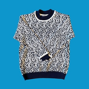 May include: A navy blue and white patterned sweater with a crew neck and long sleeves. The sweater features a repeating geometric design. The cuffs, collar, and hem are navy blue. The background is a solid blue.