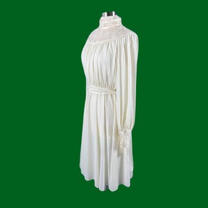 Pode incluir: Um vestido branco de manga comprida com gola alta e acabamento em renda. O vestido tem um corpete justo e uma saia fluida.