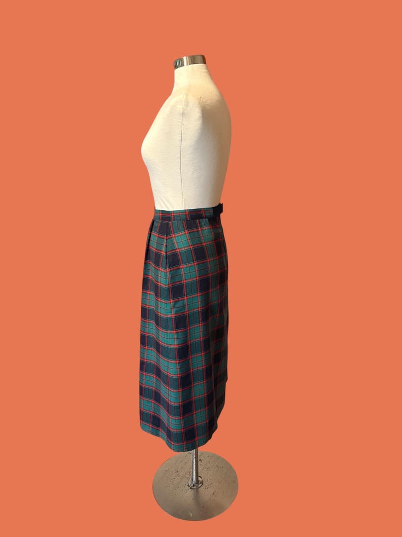 May include: A vintage plaid skirt in shades of navy, green, and red, displayed on a mannequin. The skirt features a classic plaid pattern and a tailored silhouette. The skirt is knee-length and has a belt detail at the waist.
