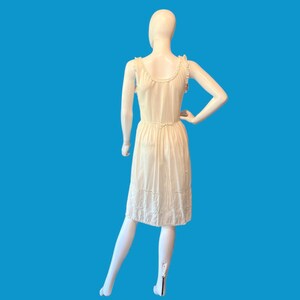 May include: A cream-colored vintage dress on a mannequin. The dress features a delicate, embroidered hemline and a cinched waist. The dress has a simple, elegant design with ruffled detailing on the shoulders and neckline. The background is a solid, bright blue.