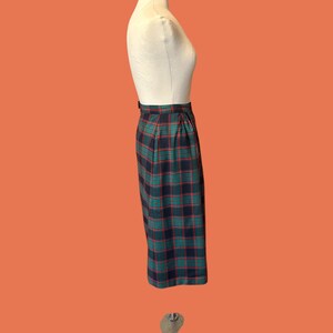 May include: A knee-length plaid skirt in shades of navy, green, and red, displayed on a mannequin. The skirt features a classic plaid pattern and a tailored fit. The garment is suspended against an orange background.