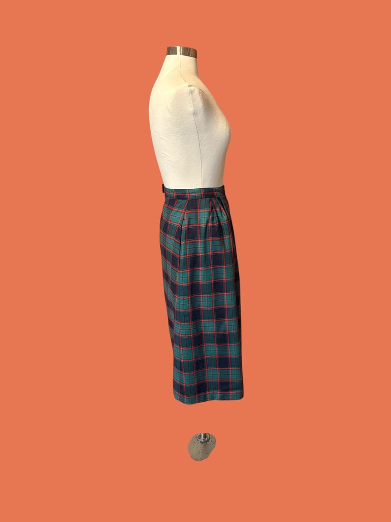 May include: A knee-length plaid skirt in shades of navy, green, and red, displayed on a mannequin. The skirt features a classic plaid pattern and a tailored fit. The garment is suspended against an orange background.