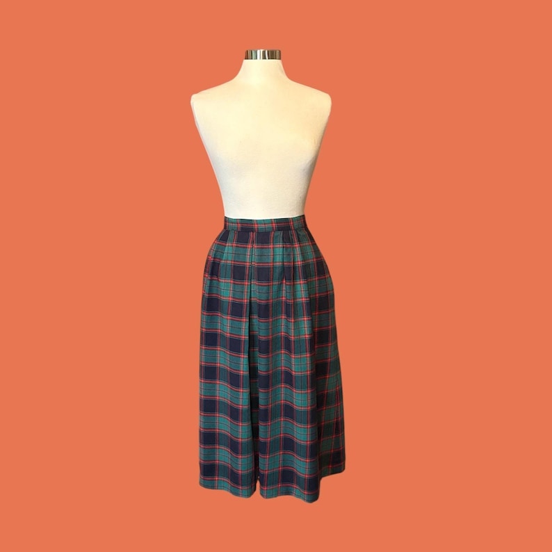 May include: A vintage plaid skirt in shades of green, navy, and red, displayed on a mannequin. The skirt features a pleated design and a high waist. The background is a solid orange color.