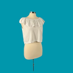 May include: White crop top with a ruffled neckline. The top is on a white mannequin.