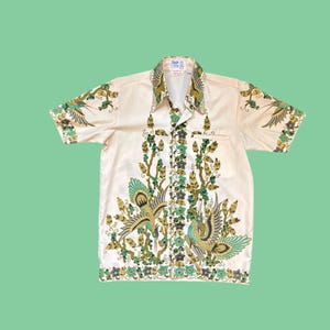 May include: A cream-coloured short-sleeved shirt with a striking bird and floral pattern in teal, green, and gold. The shirt has a classic collar and a single chest pocket. The design is repeated on the sleeves and front.