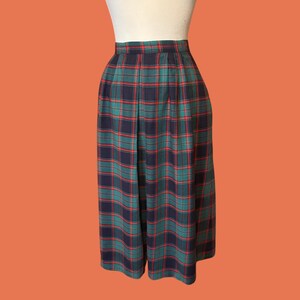May include: A pair of pleated culottes in a classic plaid pattern. The culottes feature a navy blue, green, and red tartan design. The garment is displayed on a mannequin against an orange background. The culottes are knee-length.