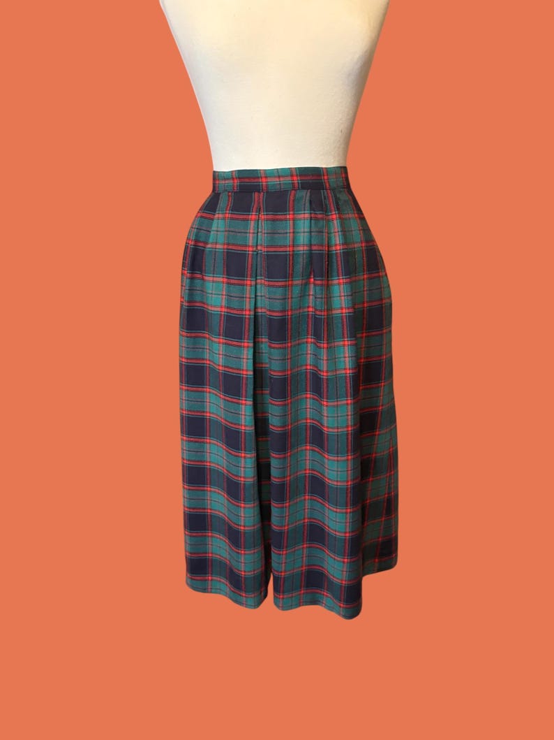 May include: A pair of pleated culottes in a classic plaid pattern. The culottes feature a navy blue, green, and red tartan design. The garment is displayed on a mannequin against an orange background. The culottes are knee-length.