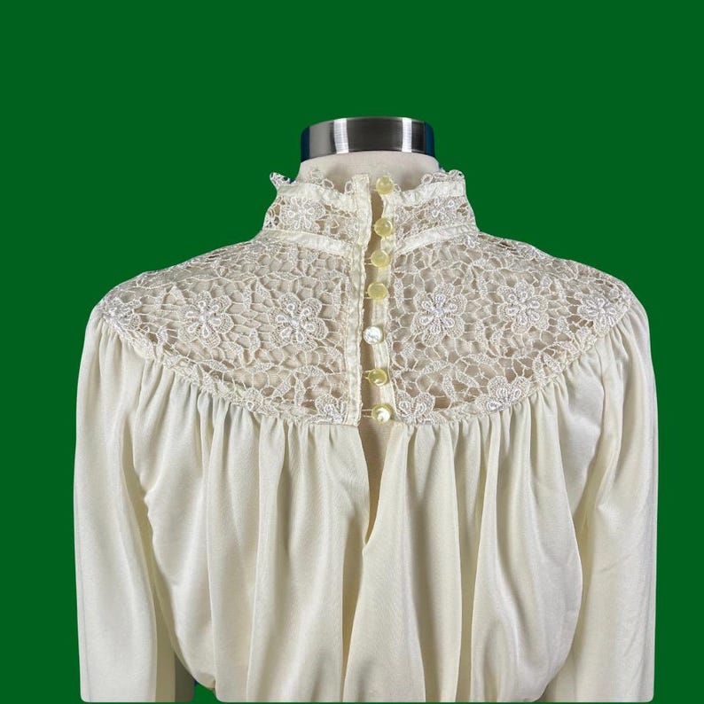 Pode incluir: Uma blusa branca de manga comprida com gola alta e uma sobreposi&ccedil;&atilde;o de renda. A renda tem um padr&atilde;o floral e &eacute; acentuada com pequenos bot&otilde;es brancos no centro das costas.