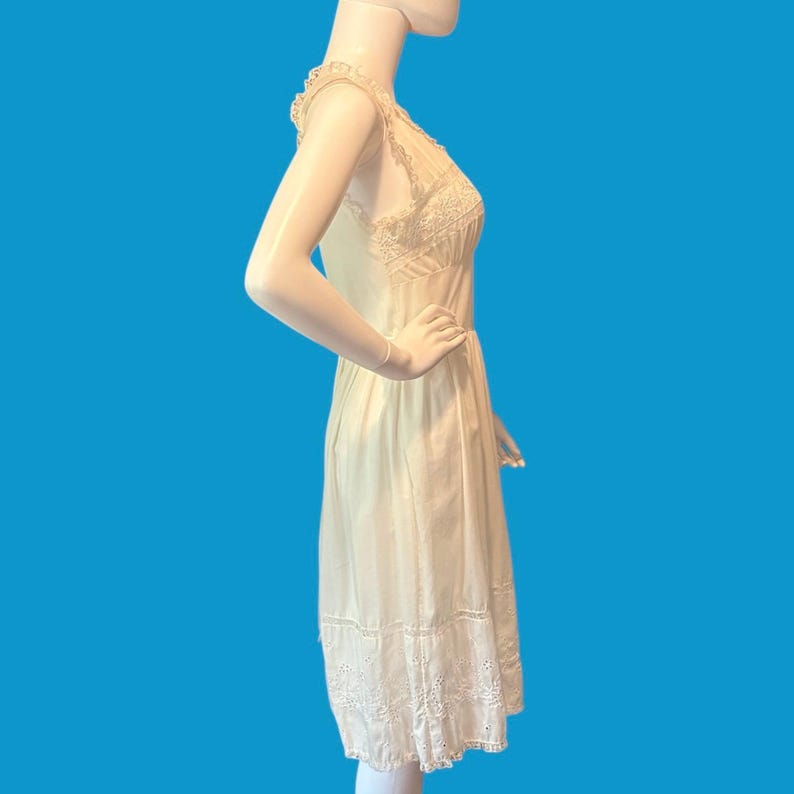May include: A cream-colored sleeveless dress with lace detailing. The dress features a scalloped neckline, lace bodice, and a hemline with embroidered cutouts. The dress is displayed on a mannequin against a blue background.