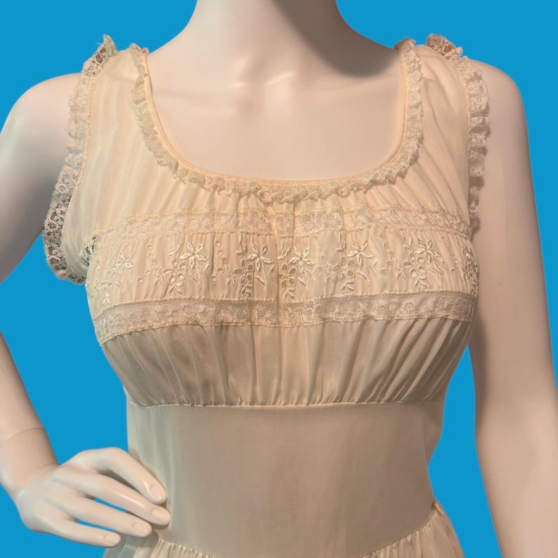 May include: Cream-colored vintage nightgown with delicate lace trim and floral embroidery. The sleeveless gown features a scoop neckline and a fitted bodice with horizontal gathering. The fabric appears lightweight and semi-sheer, with a subtle sheen.
