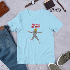 May include: Light blue t-shirt with a cartoon illustration of a man with blonde hair wearing a gray suit and jumping. The text "Pee-wee He-Man" is printed in red above the illustration.