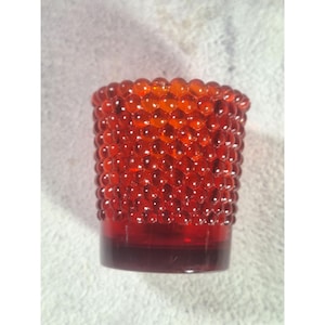 May include: A small, cylindrical, red glass tumbler with a textured, hobnail pattern. The glass is a deep red at the base, transitioning to a brighter, orange-red towards the top. The hobnail design covers the entire surface.