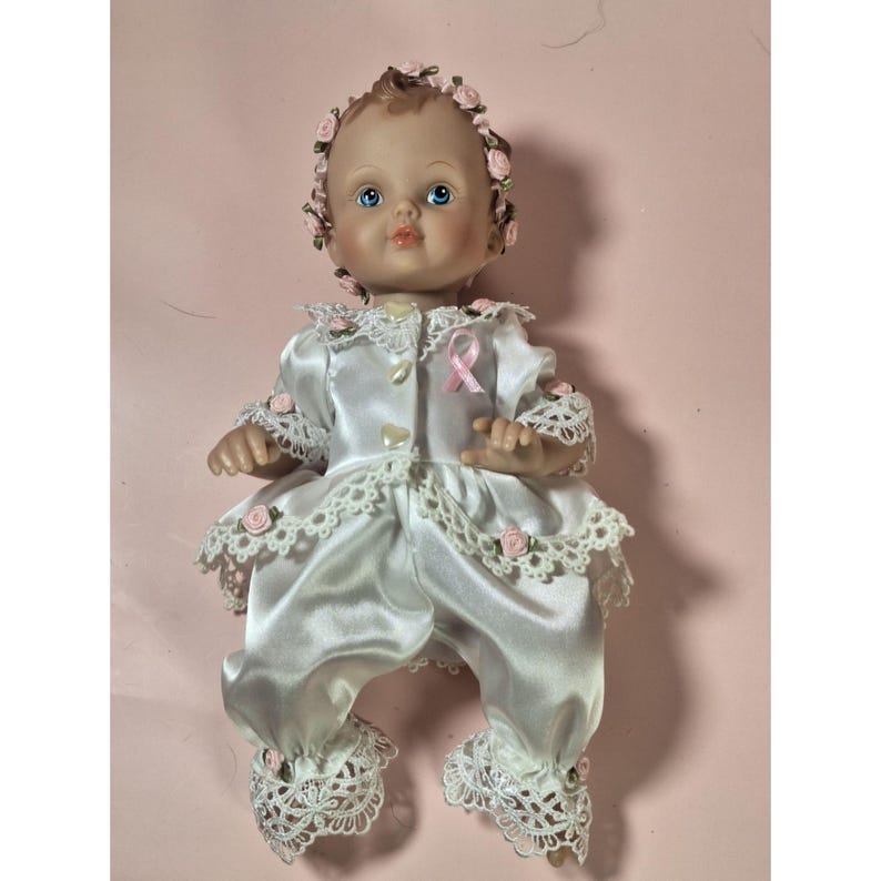 May include: A porcelain baby doll wearing a white satin dress with lace trim and a pink ribbon. The doll has a pink flower crown and blue eyes.