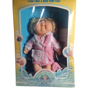 Può includere: Bambola Cabbage Patch Kids Splashin' Kids nella sua confezione originale. La bambola ha i capelli biondi e indossa un accappatoio rosa con finiture bianche. La scatola ha uno sfondo blu con grafica acquatica e la scritta "I Can Take a Bath With You!"