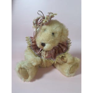 May include: A plush teddy bear with a pink and gold ruffled collar and a pink and gold bow on its head. The bear is sitting on a pink background.