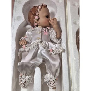 May include: A porcelain doll wearing a white satin dress with lace trim and pink rose accents. The doll has brown hair and blue eyes and is wearing a pink ribbon around her neck.