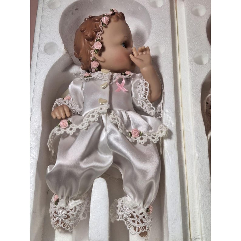 May include: A porcelain doll wearing a white satin dress with lace trim and pink rose accents. The doll has brown hair and blue eyes and is wearing a pink ribbon around her neck.