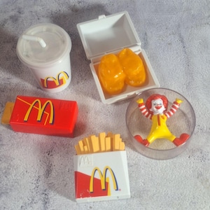 May include: A collection of vintage McDonald's Happy Meal toys. Includes a clear plastic cup with a straw, a red and yellow box, a white box with two orange nuggets, a box of fries, and a Ronald McDonald figure in a clear plastic container.