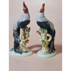 May include: A pair of ceramic rooster figurines. Each rooster has a red comb, white head and chest, and black and gray body. They perch on a tree branch with green leaves and small orange berries. The base is a light cream color.