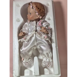 May include: A porcelain doll wearing a white satin dress with lace trim and a pink floral headband. The doll is in a white box with foam padding.