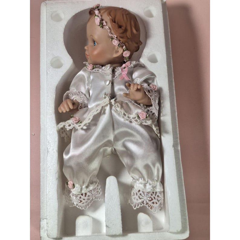 May include: A porcelain doll wearing a white satin dress with lace trim and a pink floral headband. The doll is in a white box with foam padding.