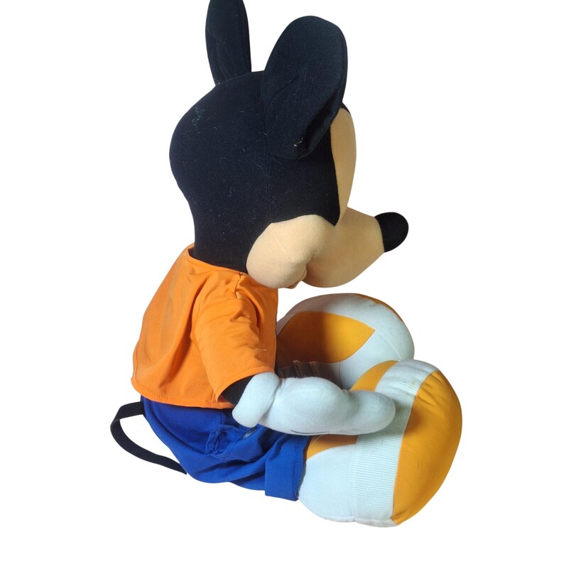 Pu&ograve; includere: Un pupazzo di peluche di Topolino, con testa nera e grandi orecchie, viso beige e naso nero. Il pupazzo indossa una maglietta arancione, pantaloncini blu e guanti bianchi. Le gambe sono bianche con strisce arancioni. Il pupazzo &egrave; seduto.