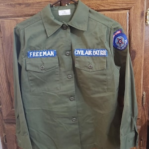 May include: A green long-sleeved shirt with a blue patch that says "CIVIL AIR PATROL" and a blue patch that says "FREEMAN". The shirt also has a blue patch with a star and the words "CIVIL AIR PATROL OKLAHOMA".