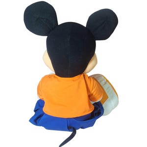 Pu&ograve; includere: Una bambola di peluche di Topolino, vista da dietro, con grandi orecchie nere e una testa nera. La bambola indossa una maglietta arancione e pantaloni blu. &Egrave; visibile una coda nera. La bambola &egrave; seduta su un cuscino bianco e arancione.