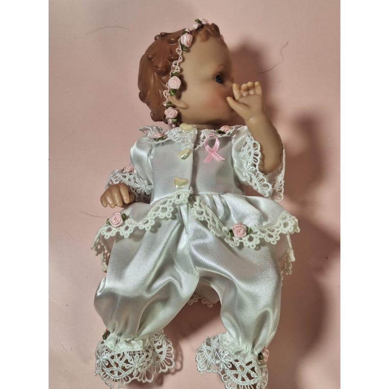 May include: A porcelain baby doll wearing a white satin dress with lace trim and pink rose accents. The doll has brown hair and is wearing a headband with pink roses.