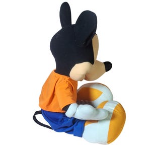 Pu&ograve; includere: Una bambola di peluche di Topolino, seduta e rivolta verso sinistra. La bambola ha una testa nera con grandi orecchie, un viso beige e un naso nero. Indossa una camicia arancione, pantaloni blu e guanti bianchi. Le gambe sono bianche con accenti arancioni e gialli.