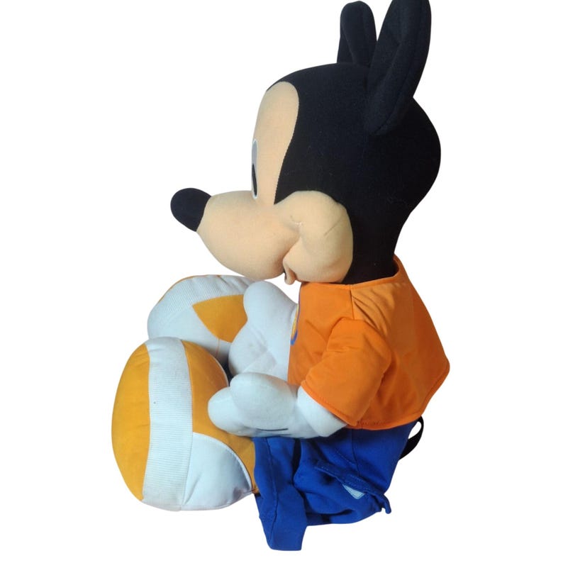 Pu&ograve; includere: Una bambola di peluche di Topolino, con una testa nera e grandi orecchie, un viso beige e un naso nero. Mickey indossa una camicia arancione, pantaloni blu e guanti bianchi. Le gambe della bambola sono bianche e gialle.