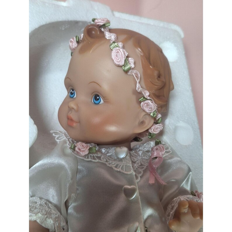 May include: A porcelain baby doll wearing a white satin dress with lace trim and a pink flower headband. The doll has blue eyes and rosy cheeks.