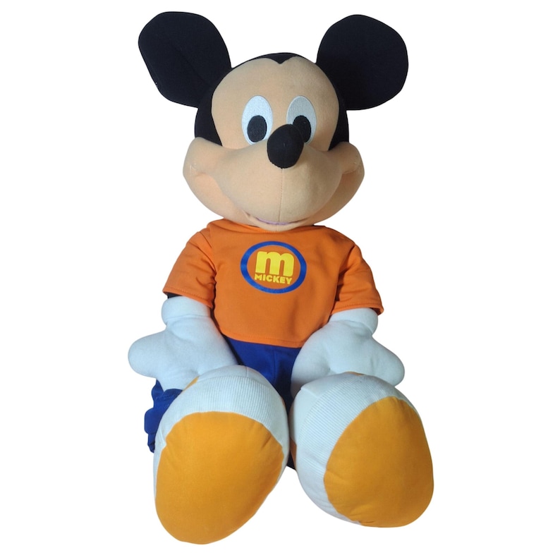 Pu&ograve; includere: Una grande bambola di peluche di Topolino. Mickey indossa una maglietta arancione con un logo "Mickey" blu e giallo, pantaloncini blu e guanti e scarpe bianche. La bambola ha orecchie nere e una faccia color camoscio.