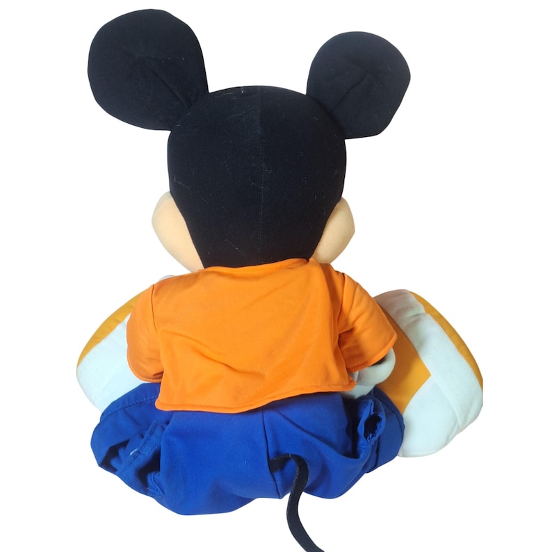 Pu&ograve; includere: Un pupazzo di peluche di Topolino, visto da dietro, con orecchie nere, testa nera e viso beige. La bambola indossa una camicia arancione e pantaloncini blu. Le scarpe sono bianche e gialle.