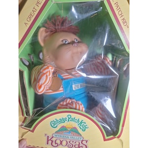 May include: A Cabbage Patch Kids Koosas doll in its original packaging. The doll has a tan face, brown hair, and is wearing blue overalls with orange and white striped sleeves. The packaging is green and yellow.