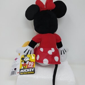 May include: A Minnie Mouse plush toy, viewed from the back, with a red dress with white polka dots, a red bow, and black ears. The toy has white gloves and a black tail. A yellow tag with Mickey Mouse and "90 Years" is attached.