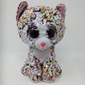 May include: A plush cat toy covered in colorful sequins. The cat has large, glittery purple eyes, pink ears, and a pink nose. The toy is white with a pink mouth and whiskers.