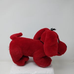 May include: A plush, red dog toy with floppy ears, a black nose, and a small black tuft of hair. The dog is in a side-facing pose, with a curled tail and a white tag visible on its side.