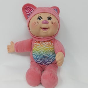 May include: A pink doll wearing a cat costume with a rainbow zebra-striped design on the front. The doll has a smiling face, brown eyes, and pink cheeks. The cat ears have a pink and purple zebra print.