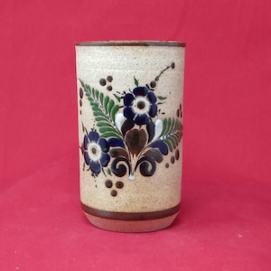 May include: A cylindrical ceramic vase with a beige speckled finish. The vase is adorned with a floral design featuring blue flowers, green leaves, and brown accents. The base and rim are a darker brown color.