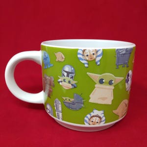 K&ouml;nnte beinhalten: Wei&szlig;er Keramikbecher mit gr&uuml;nem &Auml;u&szlig;eren, verziert mit Illustrationen von Zeichentrickfiguren. Der Becher hat ein wei&szlig;es Innenleben und einen gebogenen Griff. Die Charaktere stammen aus einem beliebten Science-Fiction-Franchise.