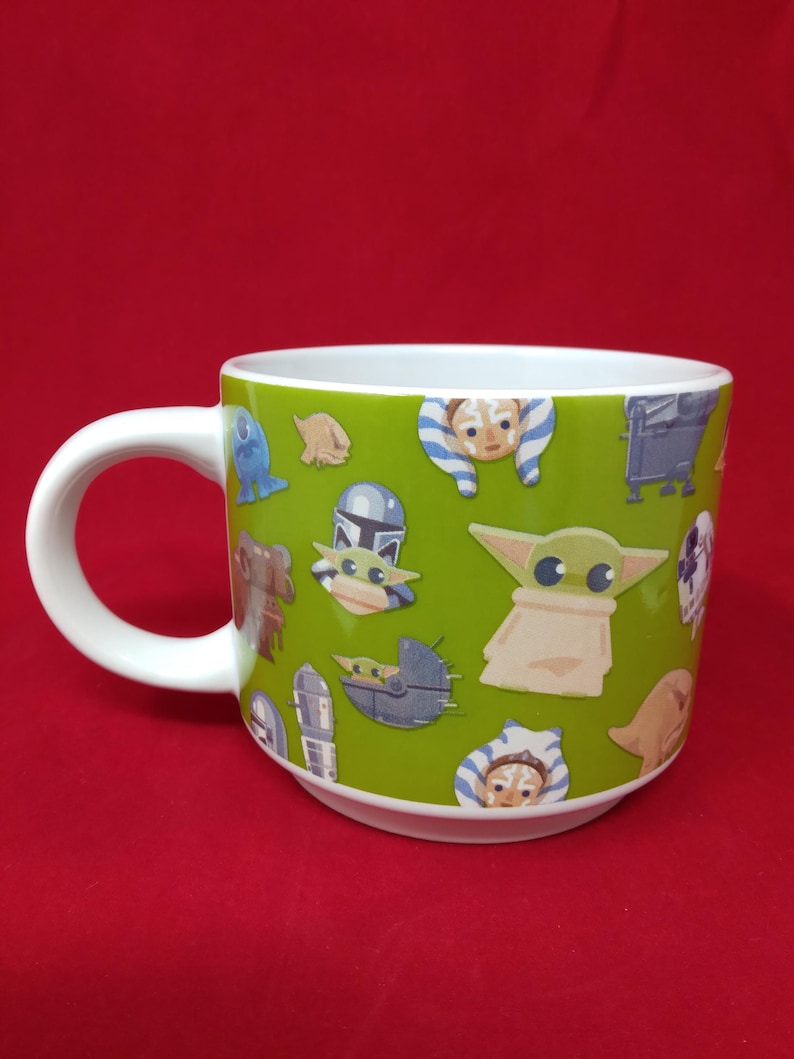 K&ouml;nnte beinhalten: Wei&szlig;er Keramikbecher mit gr&uuml;nem &Auml;u&szlig;eren, verziert mit Illustrationen von Zeichentrickfiguren. Der Becher hat ein wei&szlig;es Innenleben und einen gebogenen Griff. Die Charaktere stammen aus einem beliebten Science-Fiction-Franchise.