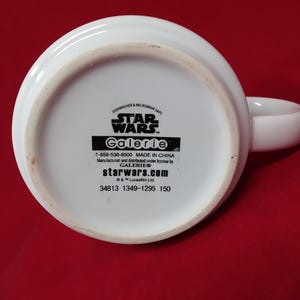 K&ouml;nnte beinhalten: Wei&szlig;er Star Wars-Becher aus Keramik mit dem Text "STAR WARS" und "Galerie" auf der Unterseite. Der Becher ist sp&uuml;lmaschinen- und mikrowellengeeignet. Der Griff ist wei&szlig; und gebogen. Auf der Unterseite des Bechers steht "starwars.com".