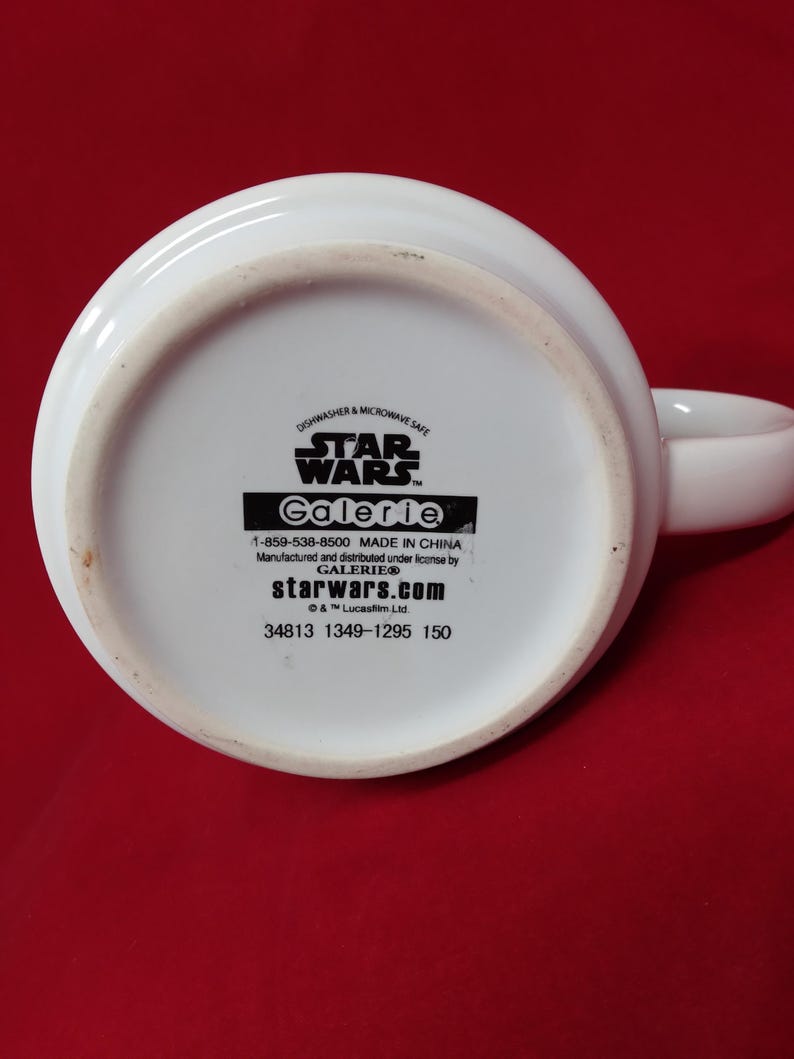 K&ouml;nnte beinhalten: Wei&szlig;er Star Wars-Becher aus Keramik mit dem Text "STAR WARS" und "Galerie" auf der Unterseite. Der Becher ist sp&uuml;lmaschinen- und mikrowellengeeignet. Der Griff ist wei&szlig; und gebogen. Auf der Unterseite des Bechers steht "starwars.com".