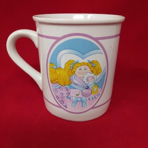 May include: White ceramic mug featuring a cartoon illustration of a blonde-haired doll in a blue heart-shaped frame. The doll is wearing a robe and holding a powder puff. The mug has a white handle and a pink and purple border.