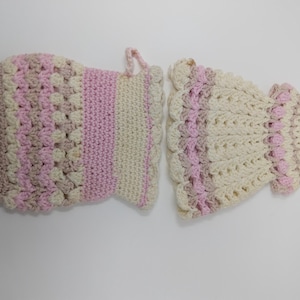 May include: Two handmade crochet pot holders in a light color palette. One is rectangular with a loop, the other is shaped like a dress. Both feature alternating stripes of cream, pink, and tan.
