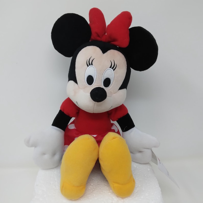 May include: A plush Minnie Mouse doll, featuring a red dress with white polka dots, black ears, and a red bow. The doll has white gloves and yellow shoes. The doll is sitting on a white surface.