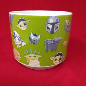 K&ouml;nnte beinhalten: Ein gr&uuml;ner Keramik-Blumentopf mit verschiedenen Star Wars-Charakteren, darunter The Mandalorian und Grogu. Der Topf hat eine wei&szlig;e Innenseite und einen roten Hintergrund.